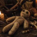 Boneco de vodu em uma mesa de madeira com velas acesas, ervas secas e contas coloridas ao redor.