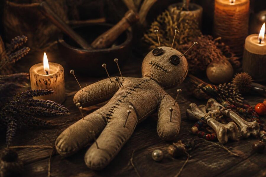 Boneco de vodu em uma mesa de madeira com velas acesas, ervas secas e contas coloridas ao redor.